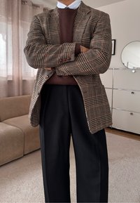 Άτομο στέκεται με τα χέρια σταυρωμένα φορώντας καφέ καρό σακάκι, μπορντό πουλόβερ, γαλάζιο πουκάμισο και μαύρο παντελόνι σε σαλόνι.