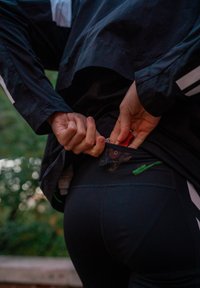 Leggings atléticas pretas com bolso traseiro com fecho, detalhe em verde e um corta-vento preto com riscas brancas. Mãos a retirar um objeto.