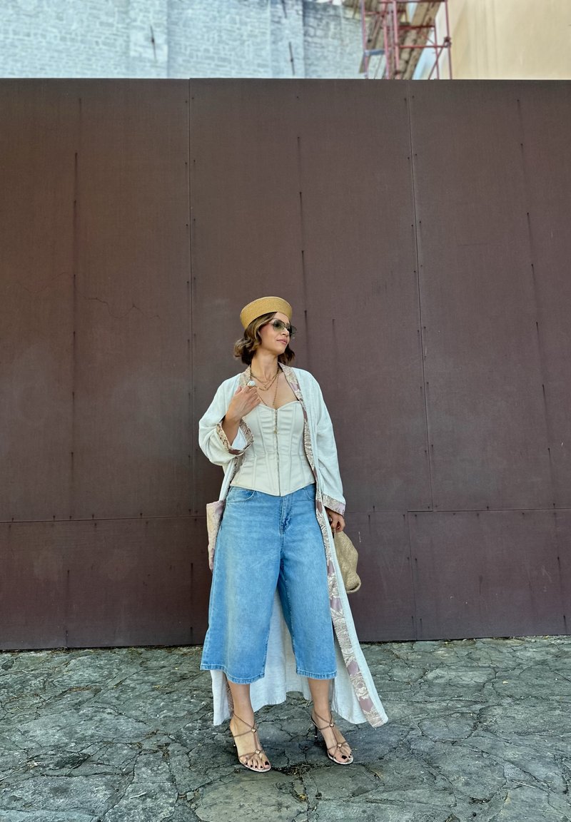 Kvinde iført brede cowboybukser, beige korset top, lang lys kappe, stråhat og solbriller, der står på revnet stensbelægning ved en brun mur.