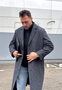 Manteau en tweed gris à la coupe structurée, fermeture par boutons, et revers en angle, associé à un pull noir et un jean décontracté.