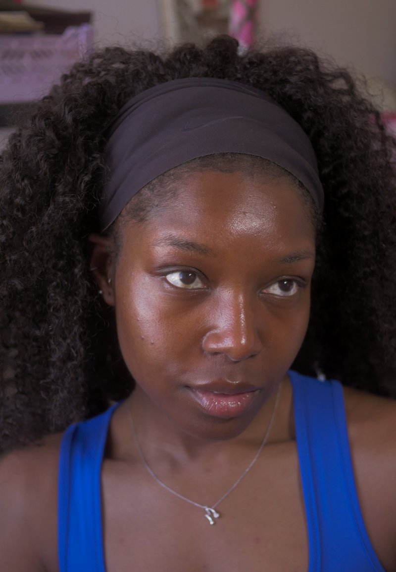 Junge Frau mit lockigem Haar, die ein schwarzes Stirnband und ein blaues Tanktop trägt, schaut leicht zur Seite mit neutralem Ausdruck.