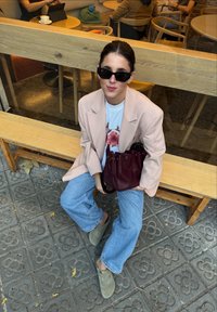 Sieviete ar saulesbrillēm, bēša žaketē, gaiši zilās džinsās, turot bordo somiņu, sēž uz koka soliņa kafejnīcā.