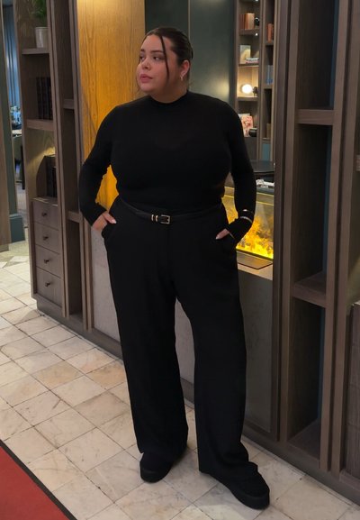 Mujer con suéter de cuello alto negro y pantalones de pierna ancha, de pie con las manos en los bolsillos junto a una chimenea moderna y una estantería.