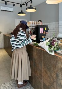 Femme en chemise rayée et jupe beige tenant une boisson froide au comptoir d'un café, avec un bouquet de fleurs et un barista en arrière-plan.