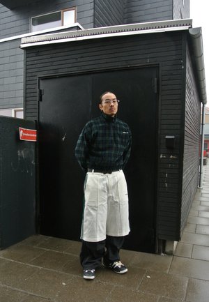 Man met een bril, een groen geruit jasje en witte Adidas-shorts over zwarte broeken staat voor een zwart gebouw met een rood sprinklerbord.