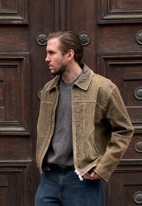 Veste en cuir marron à texture lisse, fermeture éclair et col pointu. Portée sur un pull gris, associée à un jean bleu.