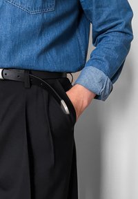 Chemise en denim (bleu) avec col à boutons, associée à un pantalon noir. Ceinture noire avec boucle métallique, une main dans la poche.