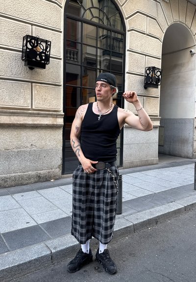 Joven con camiseta sin mangas negra, pantalones de cuadros anchos, gorra negra llevada al revés, de pie en una calle de la ciudad con tatuajes y joyas, sosteniendo una mano cerca de la cara.