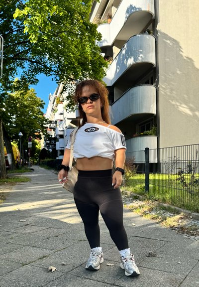 Conjunto atlético que incluye una camiseta corta blanca con logo, leggins negros de tiro alto y zapatillas plateadas. Gafas de sol y bolso de hombro beige.