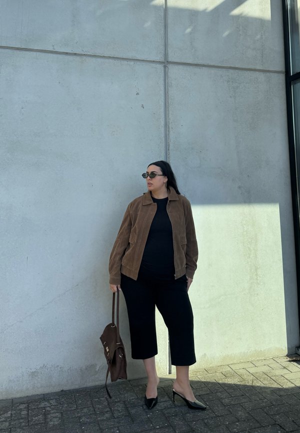 Mujer con gafas de sol, chaqueta marrón, top negro y pantalones cortos está apoyada contra una pared de cemento gris sosteniendo una bolsa marrón.
