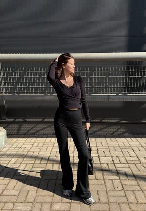 Joven mujer con top y pantalones negros de manga larga, de pie en una acera pavimentada, tocándose el cabello y sosteniendo un bolso frente a una barandilla de metal.