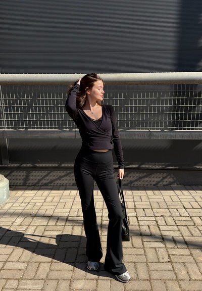 Joven mujer con top y pantalones negros de manga larga, de pie en una acera pavimentada, tocándose el cabello y sosteniendo un bolso frente a una barandilla de metal.
