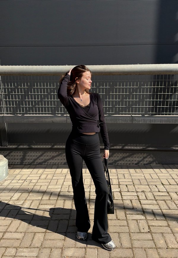 Tânără îmbrăcată într-un top negru cu mâneci lungi și pantaloni stă pe o trotuar pavat, atingându-și părul și ținând o geantă de mână în fața unui fundal cu balustradă metalică.