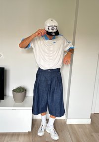 Balta trumpa rankovėmis Nike marškiniai su mėlynomis akcentais, laisvi mėlyni džinsiniai šortai, balti sportbačiai ir balta kepurė su juodu apdangalų ir grafikomis.
