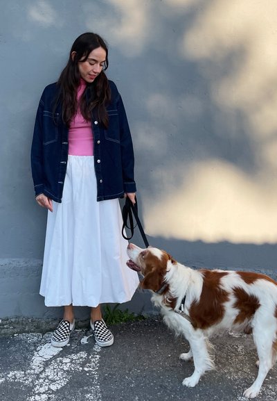 Mujer vestida con una chaqueta de mezclilla oscura, una blusa rosa y una falda blanca, de pie junto a un perro marrón y blanco con correa, frente a una pared gris.