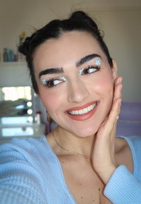 Een lachend persoon met een lichtblauwe geribbelde trui, bloeiende make-up accenten en natuurlijk ogende nagels, die hun wang vasthoudt in een heldere kamer.
