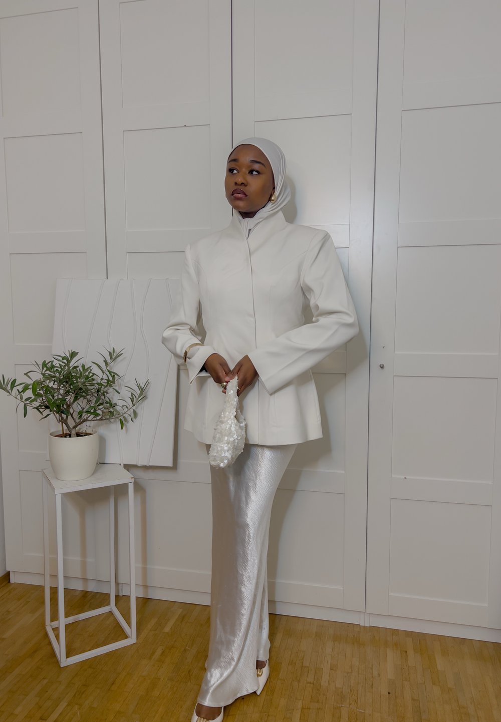 White tailored jacket with high collar, paired with a shiny silver maxi skirt. The model holds a beaded white handbag and stands beside a plant.