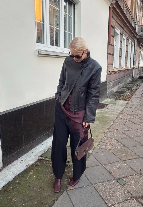 Černá kožená bunda, bordó saténový top s krajkovým lemem, široké černé kalhoty a hnědá kabelka. Bordó kotníkové boty se čtvercovou špičkou. Prostředí na chodníku.