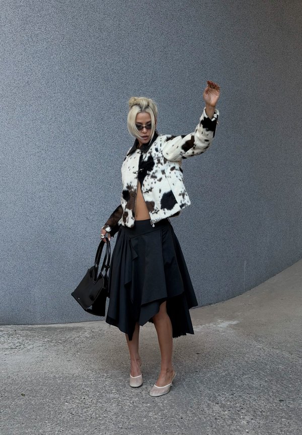 Mujer con chaqueta con patrón en blanco y negro, falda asimétrica negra, tacones y gafas de sol sostiene un bolso negro, levantando un brazo contra una pared gris.