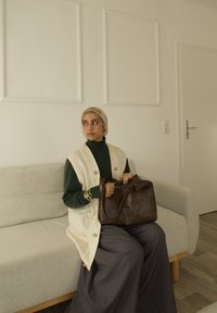 Femme portant un foulard beige et un gilet, assise sur un canapé gris, tenant un grand sac en cuir marron, regardant sur le côté dans une pièce minimaliste.