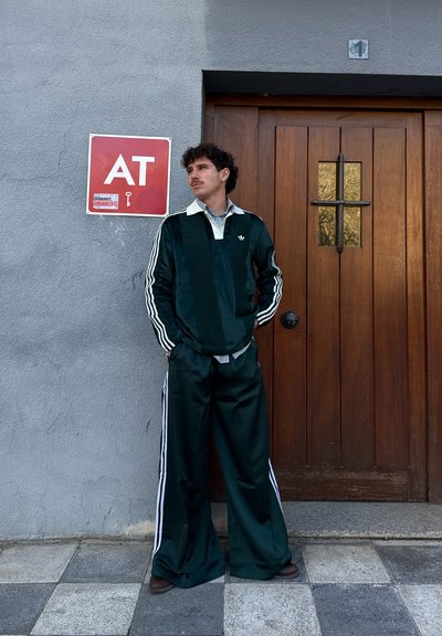 Joven con chándal Adidas verde oscuro de pie con las manos en los bolsillos cerca de una puerta de madera y un cartel rojo con "AT" en una pared gris.
