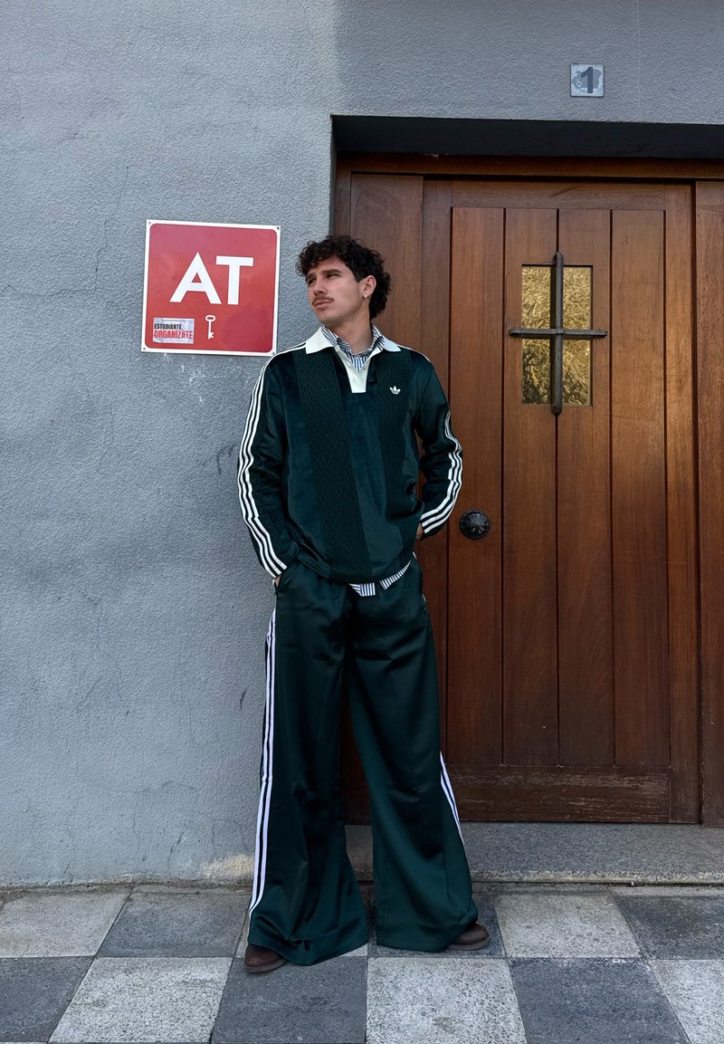 Un jeune homme en survêtement Adidas vert foncé se tient les mains dans les poches près d'une porte en bois et d'un panneau rouge avec les lettres "AT" sur un mur gris.