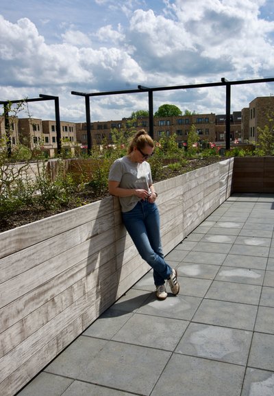 Mujer con gafas de sol apoyada en un jardinería de madera con flores en una terraza de azulejos bajo un cielo parcialmente nublado.
