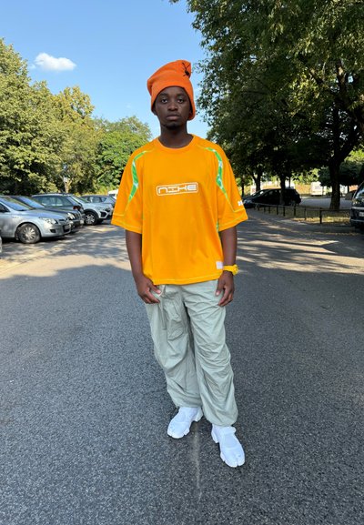 Joven de pie en una calle, vestido con un gorro naranja y una camiseta, pantalones gris claro y zapatos blancos, con coches aparcados y árboles en el fondo.