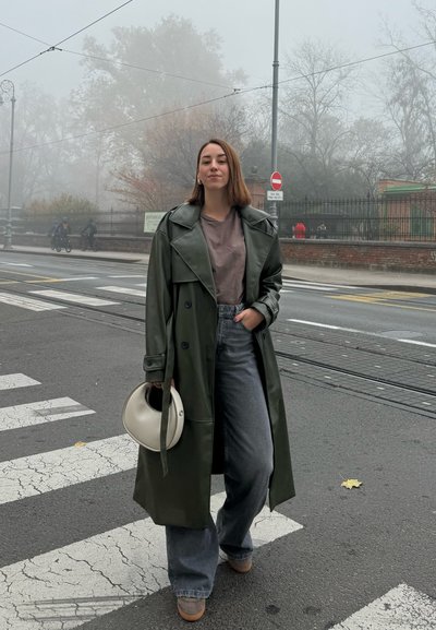 Largo abrigo de cuero verde sobre una camiseta gris, combinado con jeans gris de pierna ancha y zapatos beige, sosteniendo un bolso circular blanco.
