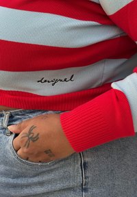 Pull côtelé rayé rouge et bleu clair avec une coupe ajustée, présentant le logo "desigual" brodé en noir sur l'ourlet. Main reposant dans une poche en denim.