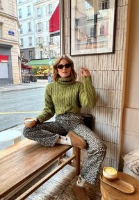 Vrouw in groene trui en panterprintbroeken, zittend met gekruiste benen op een houten bank, met een kop koffie in een café dichtbij een straatraam.