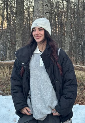 Chaqueta negra acolchada sobre un suéter gris de peluche con cuello blanco, complementada con un gorro de punto gris claro con canalé. Fondo natural al aire libre.