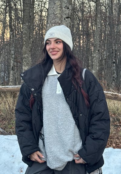 Chaqueta negra acolchada sobre un suéter gris de peluche con cuello blanco, complementada con un gorro de punto gris claro con canalé. Fondo natural al aire libre.
