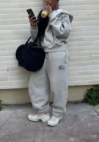 Personne portant un sweat à capuche gris clair et un pantalon de survêtement, tenant un sac de sport noir, sirotant une boisson et regardant un téléphone contre un mur carrelé.