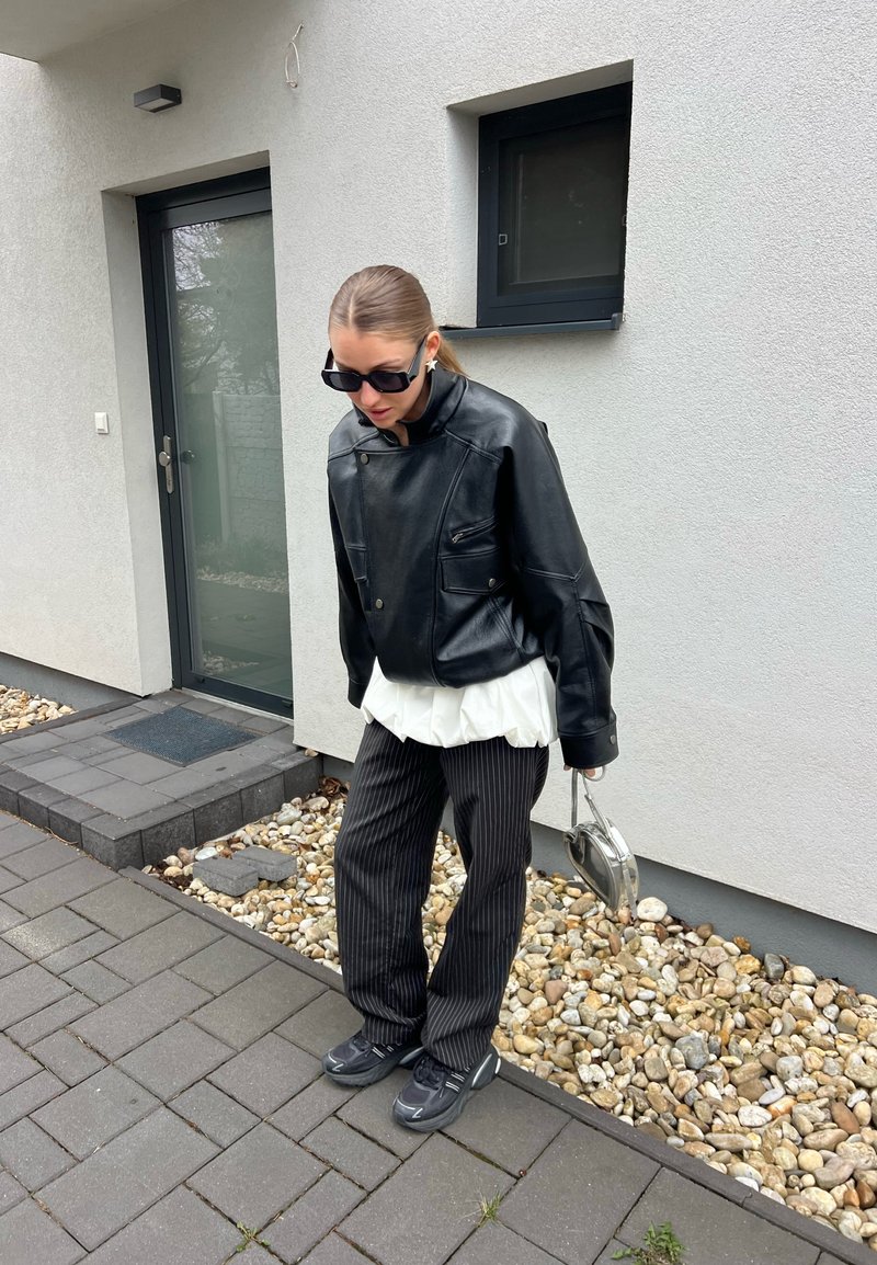 Femme portant une veste en cuir noire, un haut blanc, un pantalon à rayures fines et des lunettes de soleil, debout sur un trottoir pavé près d’un immeuble moderne gris.