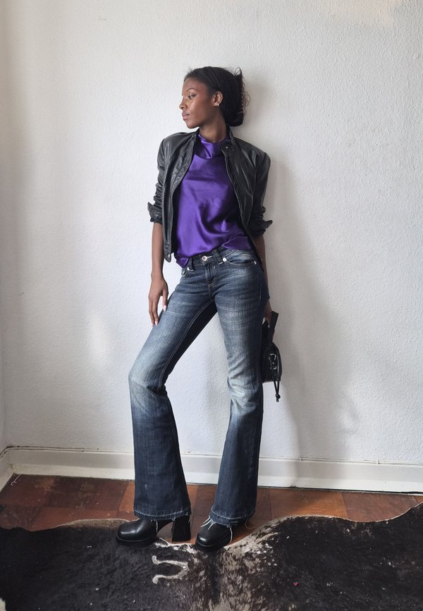 Woman in flared jeans, black leather jacket, and purple satin top stands against white wall holding a small black handbag.