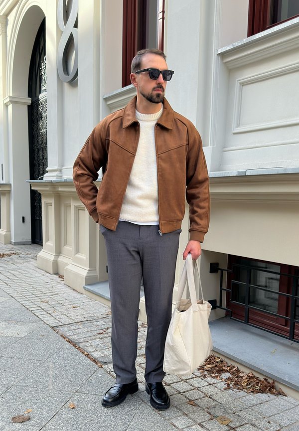 Veste en daim marron sur un pull crème, avec un pantalon gris et des chaussures noires. Tient un sac fourre-tout crème ; se tenant sur un chemin en pavés.