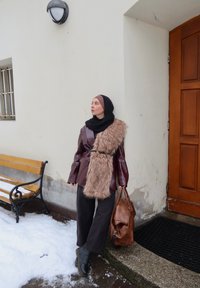 Femme portant un hijab noir, une veste en cuir marron, une écharpe en fausse fourrure et un pantalon noir, appuyée contre un mur près d'un banc enneigé et d'une porte en bois.