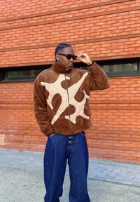 Veste marron duveteuse avec un motif abstrait crème, col montant, fermeture éclair, associée à un jean large bleu foncé, sur fond de mur en briques.