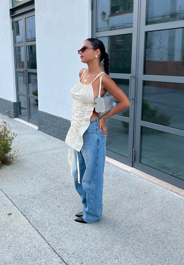 Femme aux cheveux tressés portant des lunettes de soleil, un haut crème froncé, un jean large bleu, des chaussures noires pointues et portant un sac argenté, debout devant un bâtiment moderne.