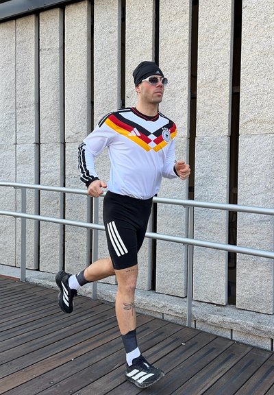 Hombre corriendo al aire libre con pantalones cortos negros, camiseta deportiva de manga larga blanca con los colores de la bandera alemana, cintillo negro, gafas de sol y zapatillas deportivas.