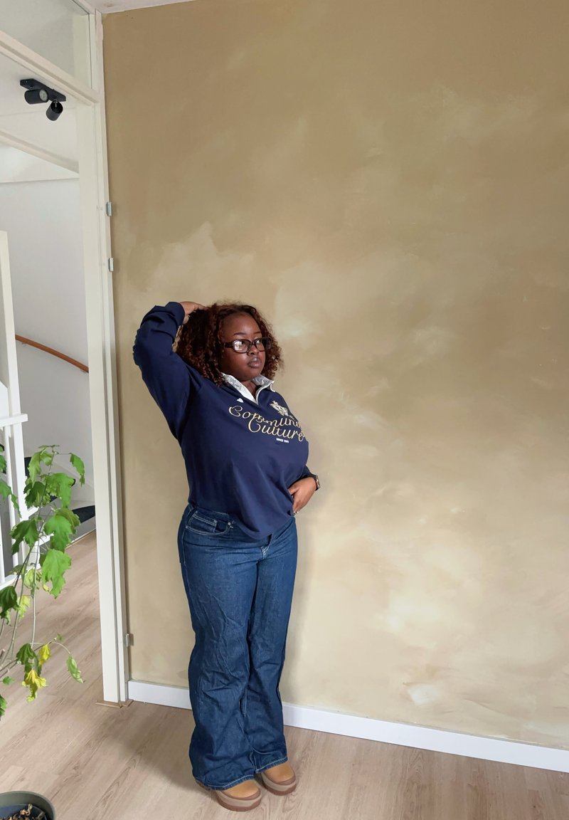 Sweatshirt azul escuro com gola branca e texto dourado, combinado com jeans azuis flare e botas castanho claro, contra uma parede bege.