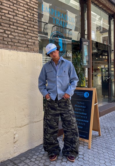 Chaqueta de lona azul con dos bolsillos frontales, combinada con pantalones holgados de camuflaje y zapatos marrones. Una gorra a rayas y gafas de sol completan el conjunto.