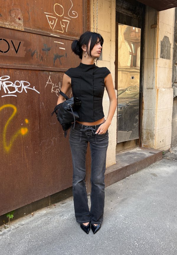 Une femme en haut noir et en jean gris se tient près d’une porte couverte de graffitis, tient un sac à main noir et regarde sur le côté.