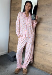 Ensemble de pyjama rose pâle avec des motifs de canards orange, manches longues et pantalon assorti. Tissu doux avec une coupe décontractée, porté avec des chaussons beige.