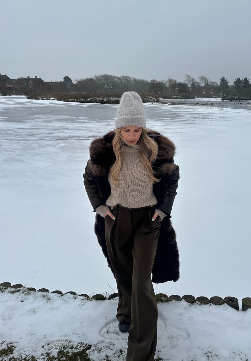 Femme portant un bonnet en tricot, un pull torsadé, un manteau en fourrure sombre et un pantalon large, debout sur un sol enneigé près d’un lac gelé avec des arbres et des maisons au loin.