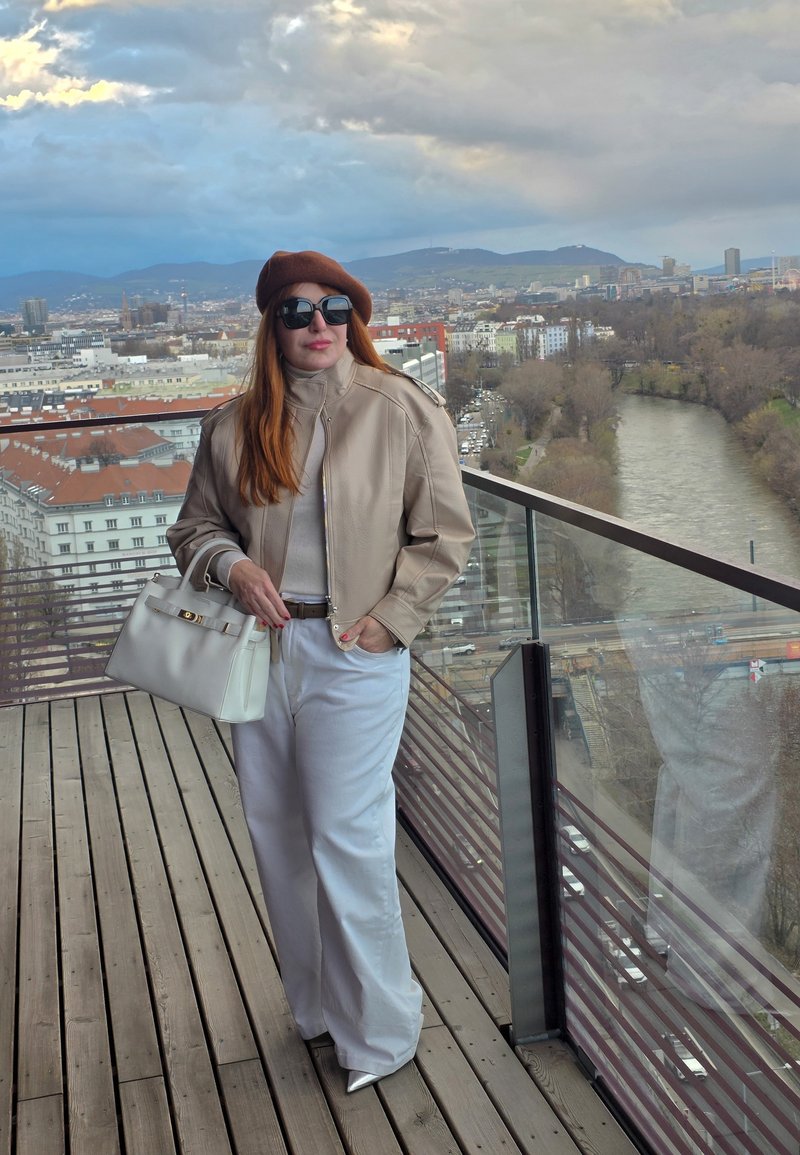 Žena v béžovém kabátu, hnědém baretě, bílých kalhotách a slunečních brýlích držící bílou kabelku na balkóně s výhledem na řeku a panorama města.