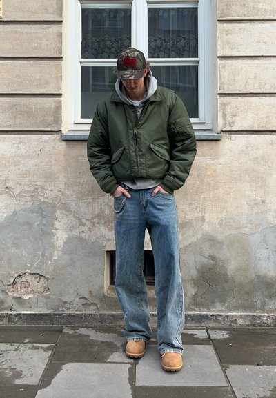 Chaqueta bomber verde, sudadera gris, jeans azul holgados con una cadena y zuecos color beige; de pie frente a una pared texturizada y una ventana.