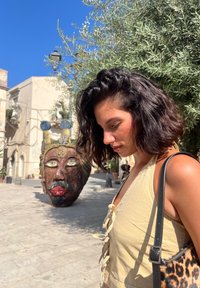 Žena s valovitom tamnom kosom nosi svijetlu žutu majicu i nosi torbu s uzorkom leoparda, stojeći blizu velikog plemenitog maske na trgu.