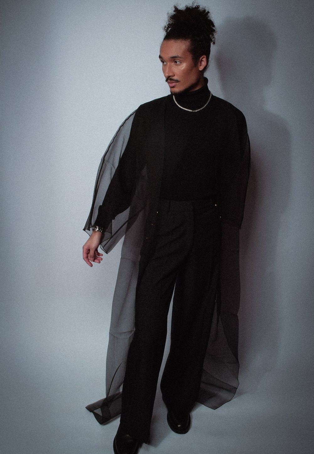 Veste kimono noire en tissu transparent par-dessus un col roulé noir et un pantalon. Manches longues, coupe ample, détails de boutons subtils. Texture lisse sur toute la surface.